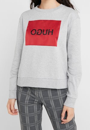 Henkilö pukeutuneena vaaleanharmaaseen collegepaitaan, jossa on punainen neliö ja musta käänteinen "HUGO"-teksti, yhdistettynä harmaisiin ja valkoisiin ruutuhousuihin.