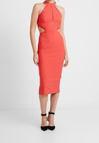 Červené midi šaty s halter výstřihem, výřezy po stranách, texturovanými krajkovými akcenty a elegantním střihem, doplněné černými botami na kotníkovém podpatku.