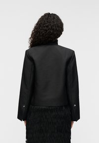 Μαύρο σακάκι με δομημένους ώμους, λείο υφάσμα και ασημένιες λεπτομέρειες στα μανίκια, συνδυασμένο με φούστα με κρόσσια σε μαύρο χρώμα.