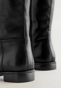 Botas de tobillo de cuero negro con un acabado liso, punta redondeada y un tacón bajo. Cuentan con un zipper lateral y costuras detalladas a lo largo de las costuras.