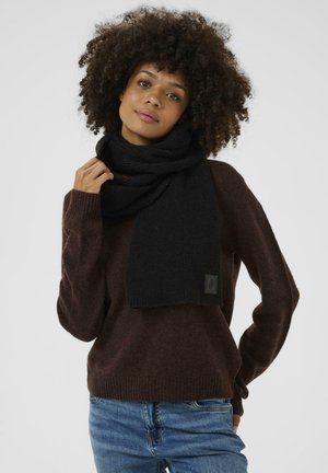 Dunkler gestrickter Schal, der um den Hals drapiert ist, aus weichem Material. Getragen mit einem braunen Pullover und blauen Jeans, mit einem Etikett auf dem Schal.