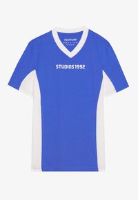 Sininen ja valkoinen lyhythihainen t-paita V-kaulaaukolla, jossa on teksti "STUDIOS 1992" valkoisena rinnassa. Silkkinen puuvillakangas.