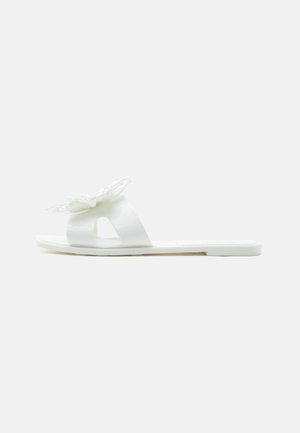 White flat slide sandal with wide strap and large layered flower decoration on top, shown in side profile on a plain background.