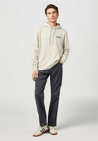 Felpa con cappuccio beige con tasca frontale e logo nero, abbinata a pantaloni grigi scuri e sneaker chiare con strisce nere.