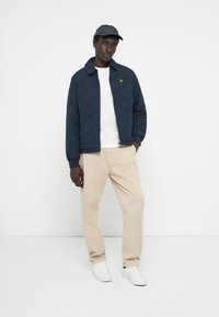 Marineblaue gesteppte Jacke mit Kragen, getragen über einem weißen T-Shirt. Beige Hosen und weiße Sneakers runden das Outfit ab.