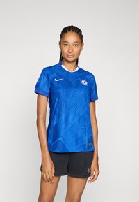 Plava Nike nogometna majica s geometrijskim uzorkom, kratkim rukavima, okruglim izrezom i Chelsea logotipom na prsima. Crne kratke hlače upotpunjuju odijelo.