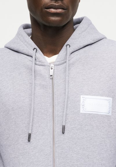 Κοντινό πλάνο ενός ατόμου που φοράει ανοιχτογκρι φούτερ με φερμουάρ και κουκούλα, με ένα λευκό ορθογώνιο αυτοκόλλητο στο στήθος.