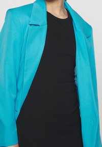 Tealová sako s hladkou texturou a strukturovanými rameny, př layeringem přes úzké černé žebrované šaty.