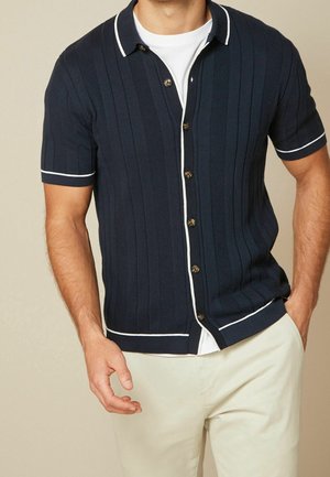 Chemise en maille à manches courtes marine avec des rayures verticales, accents blancs sur le col et l'ourlet, et boutons marron. Portez par-dessus un T-shirt blanc.