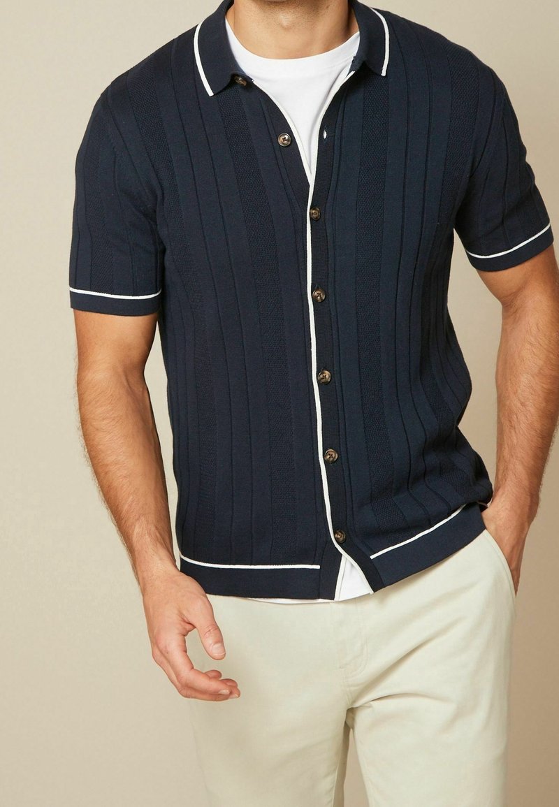 Laivastonsininen lyhythihainen neulepaita, jossa on pystysuorat raidat, valkoiset yksityiskohdat kauluksessa ja helmassa sekä ruskeat napit. Paita on puettu valkoisen T-paidan päälle.