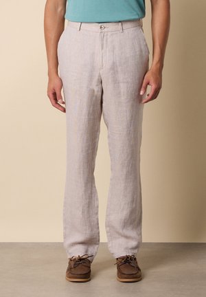 Man met beige linnen broek, ingestopt teal shirt en bruine leren mocassin schoenen staand op een grijze vloer.