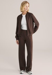 Nő barna cipzáras dzsekiben és hozzáillő nadrágban, fehér inggel alatta, világos színű tornacipőben, bézs háttér előtt áll.