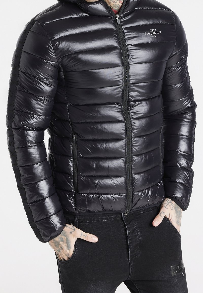 Sort dunjakke med en blank finish, der har vandrette syninger og en fuld lynlås foran. Inkluderer sidelommer og en hætte.