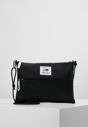 Zwarte rechthoekige crossbodytas met voorvak met rits, witte ritstrekken en een wit label met tekst aan de bovenkant voorkant.