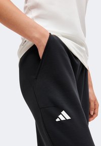 Άτομο που φοράει μαύρο παντελόνι Adidas με το χέρι στην πλαϊνή τσέπη και λευκό πουκάμισο με ορατό λογότυπο Climalite στο πλάι.