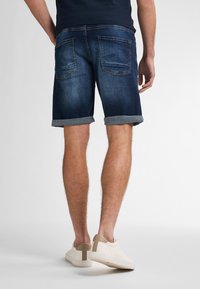 Pantalones cortos de mezclilla en azul oscuro con dobladillos enrollados. Cuenta con dos bolsillos traseros y un corte clásico. Llevados con zapatillas de color claro.