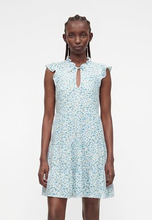 Kvinde iført en lyseblå, blomsterprintet, ærmeløs kjole med flæsekant og en lille nøglehul-udskæring, stående foran en ensfarvet hvid baggrund.