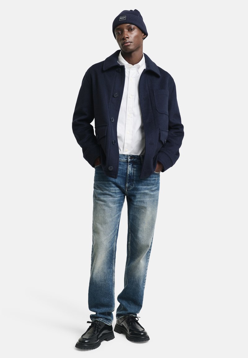Veste en mélange de laine bleu marine avec un col, fermeture à boutons et poche avant ; associée à un jean en denim bleu clair et des chaussures noires.