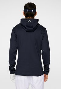 Felpa con cappuccio navy realizzata in tessuto liscio. Presenta maniche lunghe, un design aderente e un logo sul collo posteriore. Pantaloni bianchi visibili sotto.