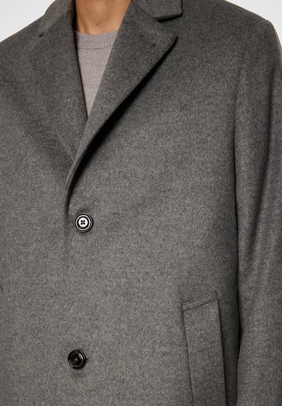 Uomo che indossa un cappotto di lana grigia con due bottoni neri e un colletto a punta sopra un maglione di lana grigia chiara.