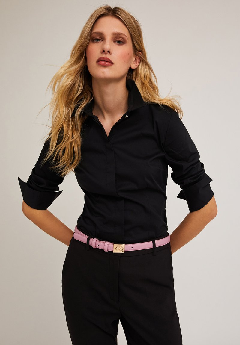 Camisa negra ajustada con mangas enrolladas, combinada con un cinturón de cuero rosa que tiene una hebilla de tono dorado. Diseño simple con un cuello clásico.