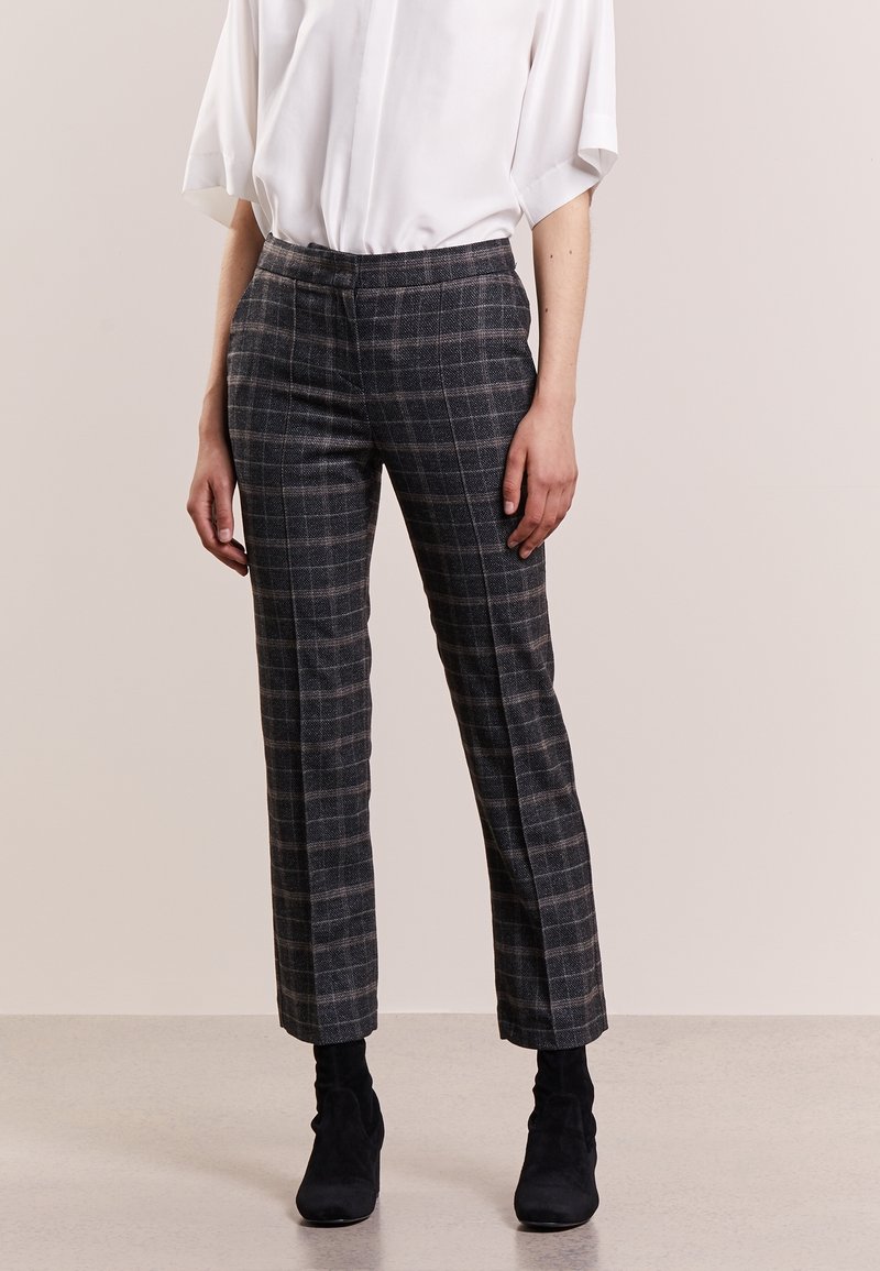 Personne portant un pantalon droit à carreaux gris, un chemisier blanc à manches courtes et des bottines noires, debout devant un fond neutre.