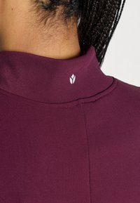 Μπορντό τοπ με ψηλό λαιμό και λεία υφή με λεπτομέρειες ραφών. Διαθέτει μικρό λευκό λογότυπο στο πίσω μέρος του λαιμού.