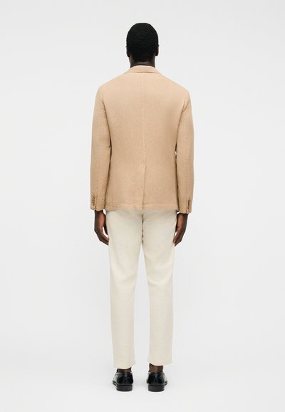 Άνδρας που στέκεται με την πλάτη προς τα εμάς, φοράει μπεζ σακάκι, εκρού παντελόνι και μαύρα παπούτσια σε λευκό απλό φόντο.