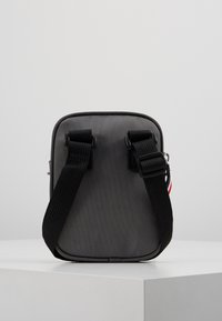 Borsa a tracolla compatta grigia con cinghia nera regolabile e un piccolo etichetta in tessuto rossa, bianca e blu sul lato, posizionata su una superficie bianca.