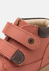 Scarpa in pelle rossa con accenti marroni, cinturino in velcro, dettagli cuciti e un logo rotondo sul lato. Fodera interna morbida visibile.