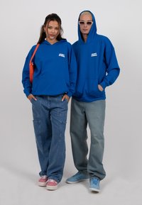 Du modeliai vilki atitinkančias mėlynas džemperius su priekiniais kišenėmis ir logotipu, derinamus su laisvais džinsais ir spalvingais sneakeriais.