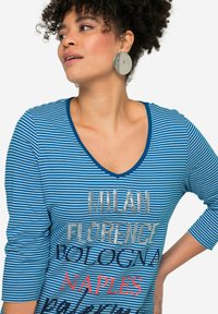 Blauw-wit gestreepte T-shirt met V-hals, voorzien van bedrukte stadsnamen in zilver en rood. Model draagt grote zilveren ronde oorbellen.