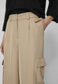 Pantalones beige con pliegues, bolsillos laterales y un detalle de solapa de carga. Fabricados en una tela suave, con una cinturilla ajustada.