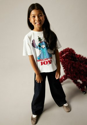 Vit t-shirt med färggrafik av Stitch-karaktären och text, ihopparad med mörka jeans och ljusbruna platta skor. Röd glitterdekoration i närheten.