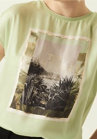Top de manga corta de color verde claro con un estampado fotográfico de hojas de palma encerradas en un marco rectangular con foil dorado. Textura suave.