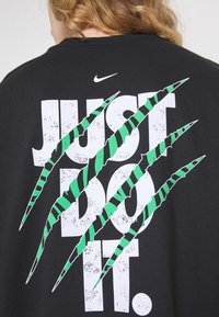 Svart T-shirt med stor vit text "JUST DO IT" och gröna klösmärken. Tillverkad i bomull med en grafisk design på baksidan.