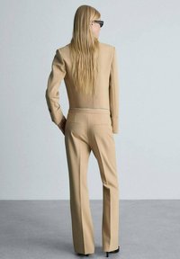 Completo beige su misura con giacca corta e pantaloni a gamba dritta. Tessuto morbido, senza motivi visibili, vista posteriore, con un accento argentato.