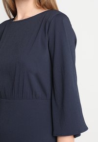 Námořnickou modrou šaty s dlouhými, rozšířenými rukávy, s kulatým výstřihem a nabíraným pasem. Vyrobeno z lehké, texturované tkaniny.