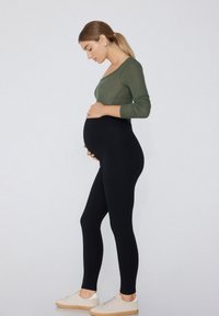 Leggings noirs en matériau extensible, associés à un haut à manches longues en olive côtelé. Le modèle caresse son ventre rond.