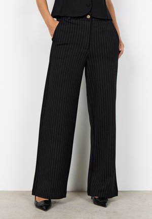 Vrouw die zwarte wijde pijpen met krijtstreep en zakken draagt, gecombineerd met zwarte schoenen met puntige neus en een zwart cropped topje ter hoogte van de taille.