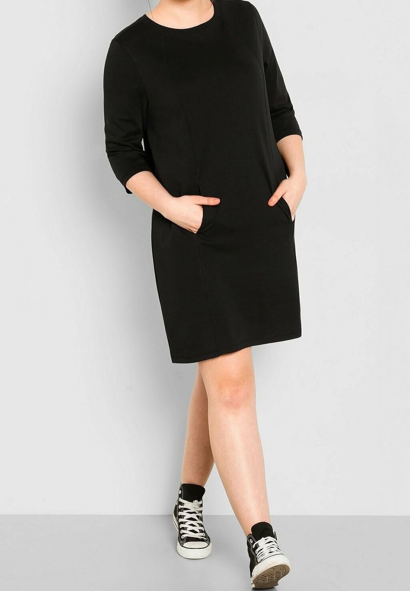 Femme portant une robe noire mi-longue aux manches trois-quarts et des baskets montantes noires avec des lacets blancs, mains dans les poches.