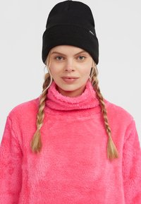 Roze fleece trui met hoge kraag en zachte textuur; gecombineerd met een zwarte gebreide muts. Heeft twee vlechten in het haar.
