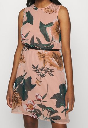 Robe rose sans manches avec de grands motifs floraux verts, blancs et marron, détail en dentelle aux épaules et une fine ceinture noire à la taille.