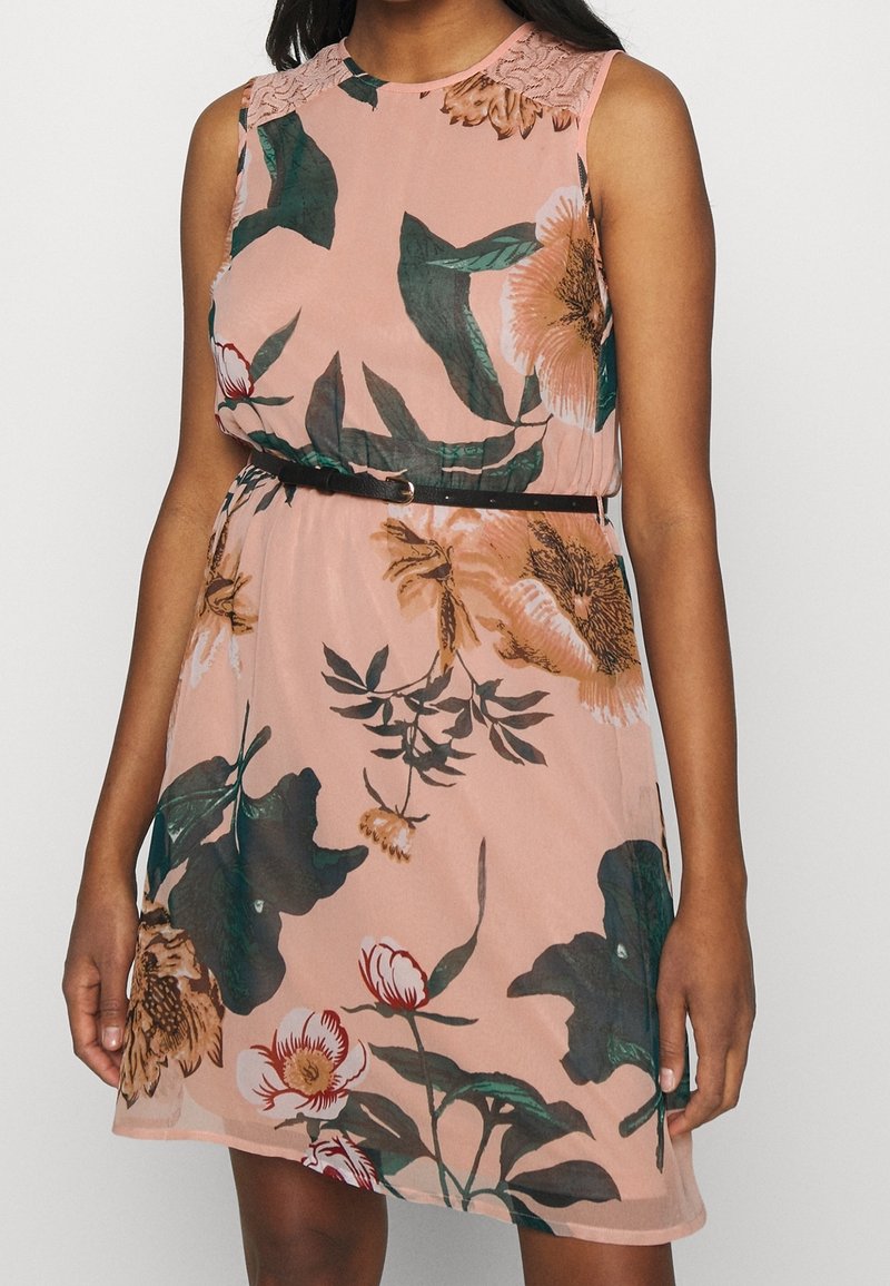 Robe rose sans manches avec de grands motifs floraux verts, blancs et marron, détail en dentelle aux épaules et une fine ceinture noire à la taille.