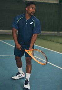 Tumesinine polo särk roheliste aktsentidega, sinised lühikesed püksid, hoiab oranži tennisereketi ja tennisepalli, jalas valged spordijalatsid ja sokid.