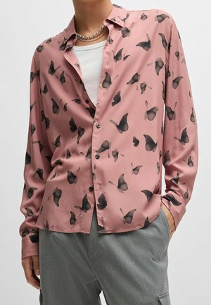 Personne portant une chemise rose à boutons avec des motifs de papillons noirs, un sous-t-shirt blanc, un pantalon gris et deux colliers en argent.