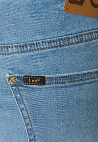 Jeans de mezclilla azul claro con un bolsillo trasero que tiene una etiqueta negra con la inscripción "Lee". Costuras amarillas visibles y un ligero detalle de desgaste.