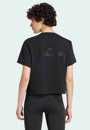 Osoba o kręconych włosach, ubrana w czarny krótki T-shirt Adidas z napisem "The Artist Athlete" i czarne legginsy, stojąca tyłem na jednolitym tle.