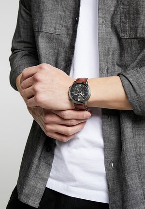 Persoon die een grijs overhemd draagt over een wit T-shirt, verstelt een polshorloge met een bruine leren band en een zwarte wijzerplaat.