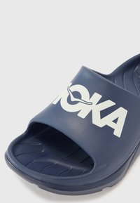 Mörkblå glidsandal i texturerad gummi, med en bred öppen tådesign och en framträdande vit logotyp över den övre remmen.